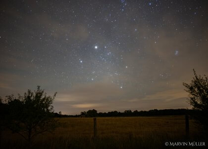 Sternenhimmel über einem Feld: Im Vordergrund sind ein Zaun sowie Büsche zu erkennen, im Hintergrund ein Feld sowie einige Bäume. Darüber leuchten die Sterne, die an einigen Stellen von Wolken verdeckt werden.