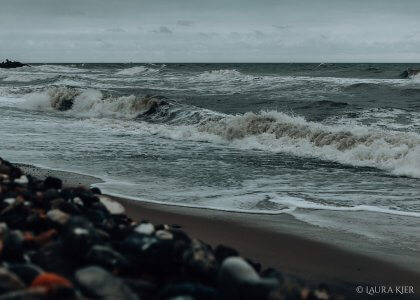 stürmische Nordsee - im Vordergrund sind Steine erkennbar, dahinter brechen sich wild schäumend die Wellen am Strand. Der Himmel ist grau und von Wolken verhangen.